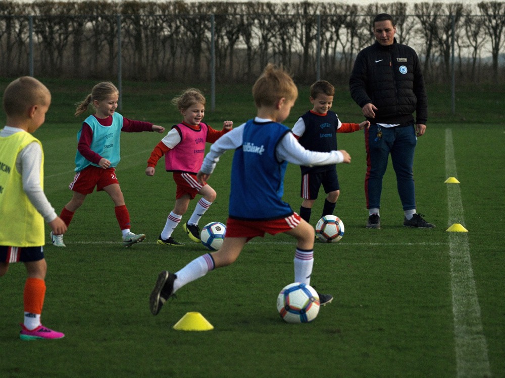 Des enfants en chasubles de couleur conduisent le ballon entre des plots, sous l'œil de leur éducateur.