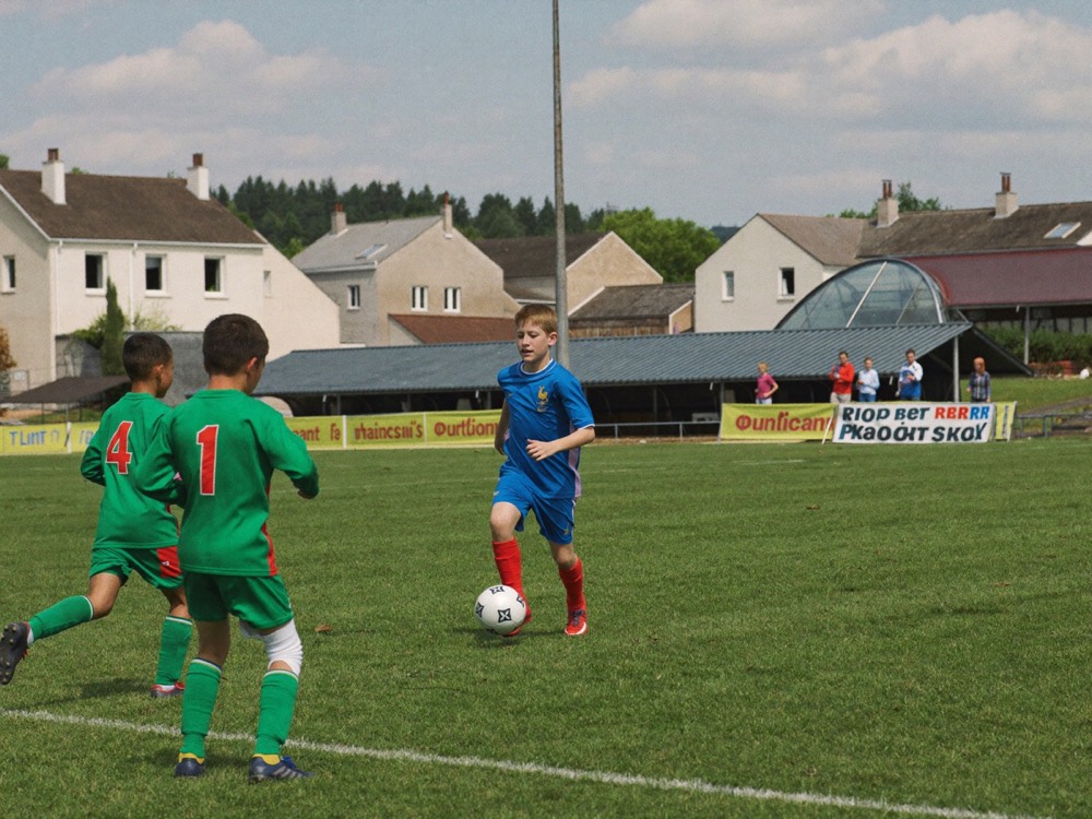 Un jeune joueur en maillot bleu s'apprête à frapper, face à deux adversaires en vert.
